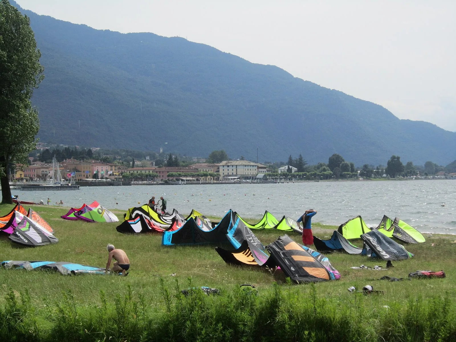 Vele da kitesurf colorate stese sul prato in riva al lago a Colico, con alcuni praticanti che preparano l'attrezzatura, il lago increspato dal vento e sullo sfondo il paese ai piedi delle montagne