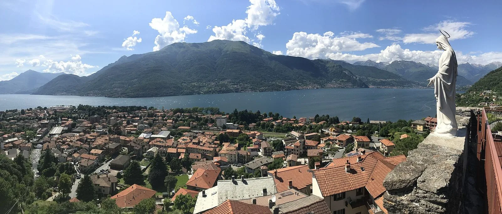 Panorama dall'alto su Dervio: i tetti rossi del paese, il lago di Como blu e le montagne della sponda opposta, con una statua bianca della Madonna sul parapetto in pietra della torre a destra