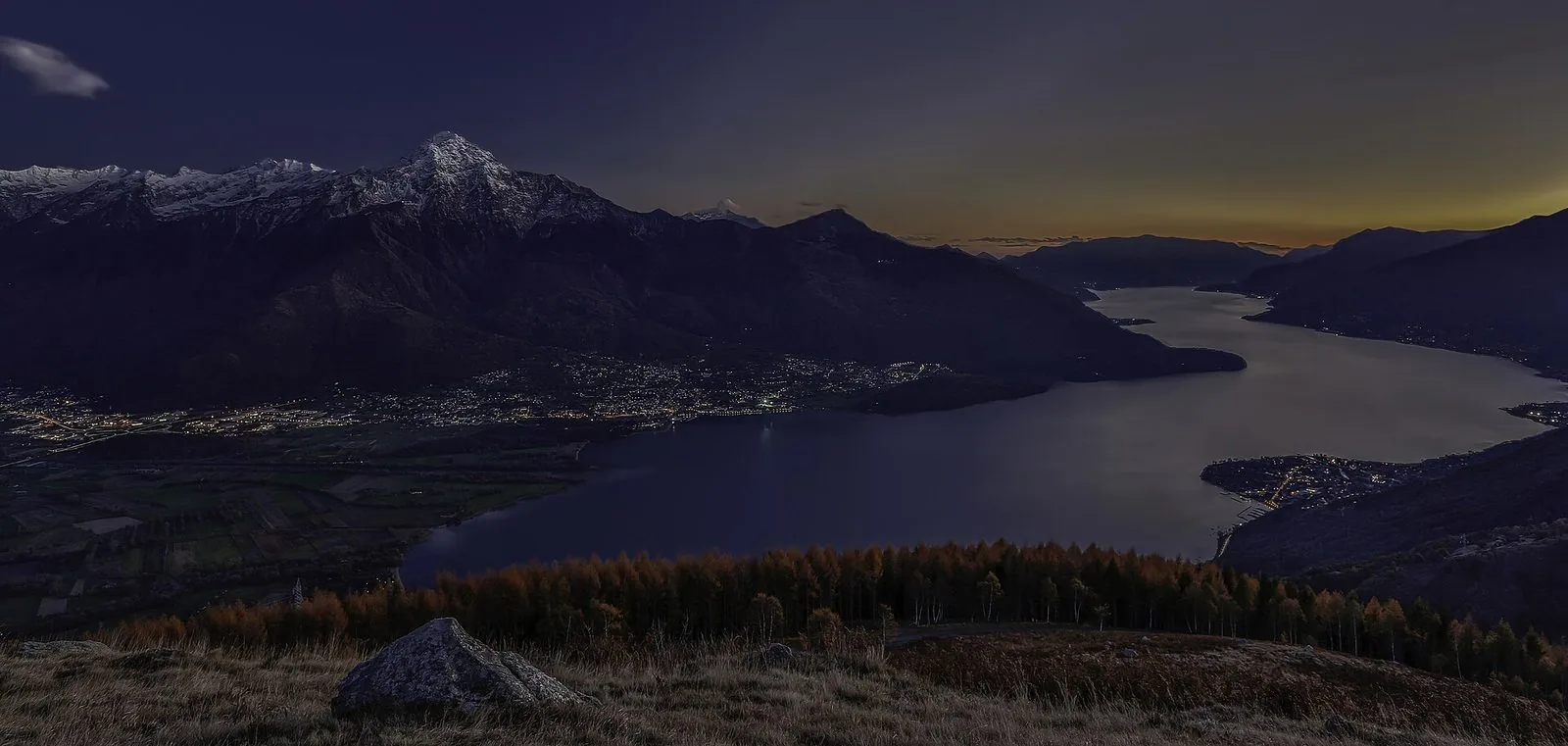 Veduta dall'alto dell'Alto Lario al crepuscolo in autunno: in primo piano un prato con un masso e un bosco di larici arancioni, sotto il lago scuro con le luci dei paesi sulla piana e a sinistra le montagne con la vetta innevata