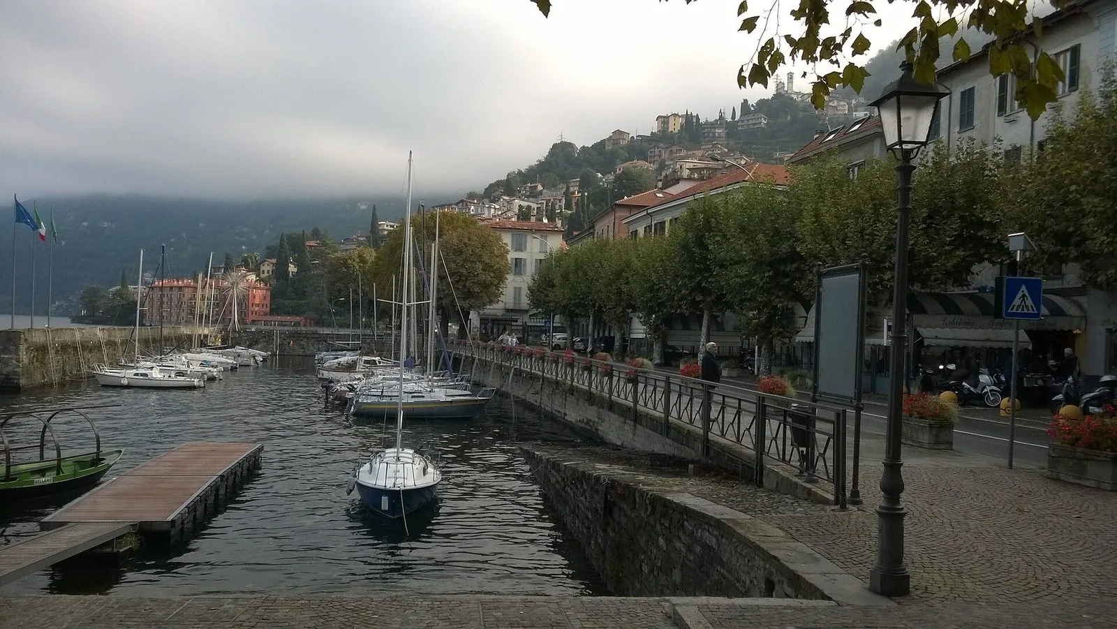 Il porticciolo di Bellano in una giornata nuvolosa, con barche a vela ormeggiate, una tradizionale barca da lago verde in primo piano a sinistra e il lungolago alberato con le case che salgono sulla collina