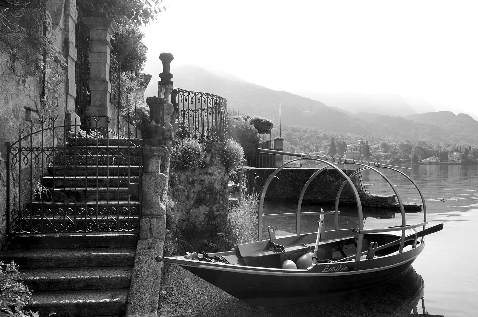 Fotografia in bianco e nero di una Lucia, la tradizionale barca da pesca del Lago di Como con i tre archi in legno, ormeggiata ai piedi di una scalinata in pietra con cancello in ferro battuto sulla riva a Mandello del Lario