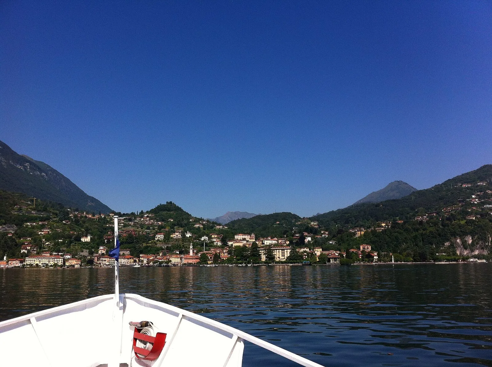 La prua bianca di un battello con il tubo antincendio rosso avanza sull'acqua verso un paese della riva occidentale del Lago di Como, con case colorate, un campanile e colline verdi sotto un cielo blu terso