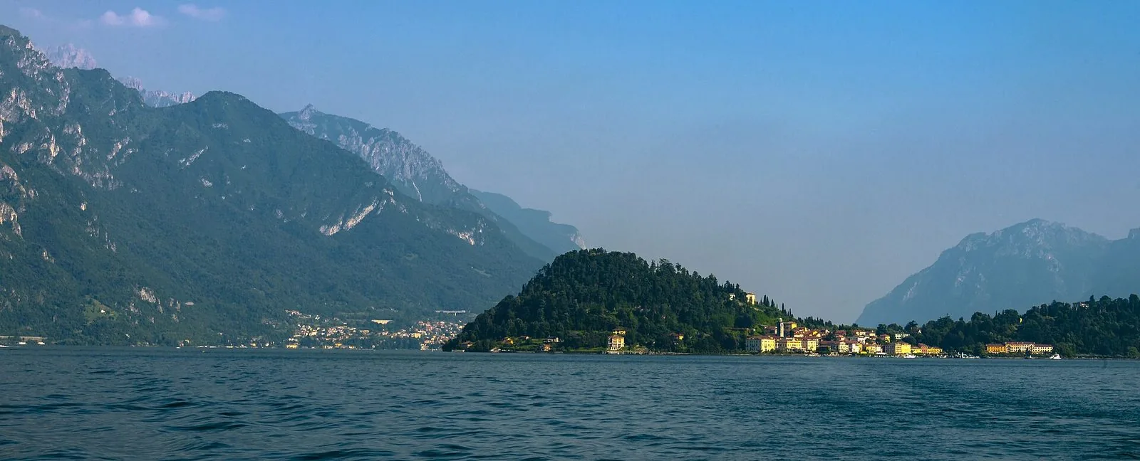 Bellagio vista dall'acqua durante la traversata: il promontorio boscoso con le case gialle e arancioni del borgo sulla riva, il lago increspato in primo piano e le montagne nella foschia sullo sfondo