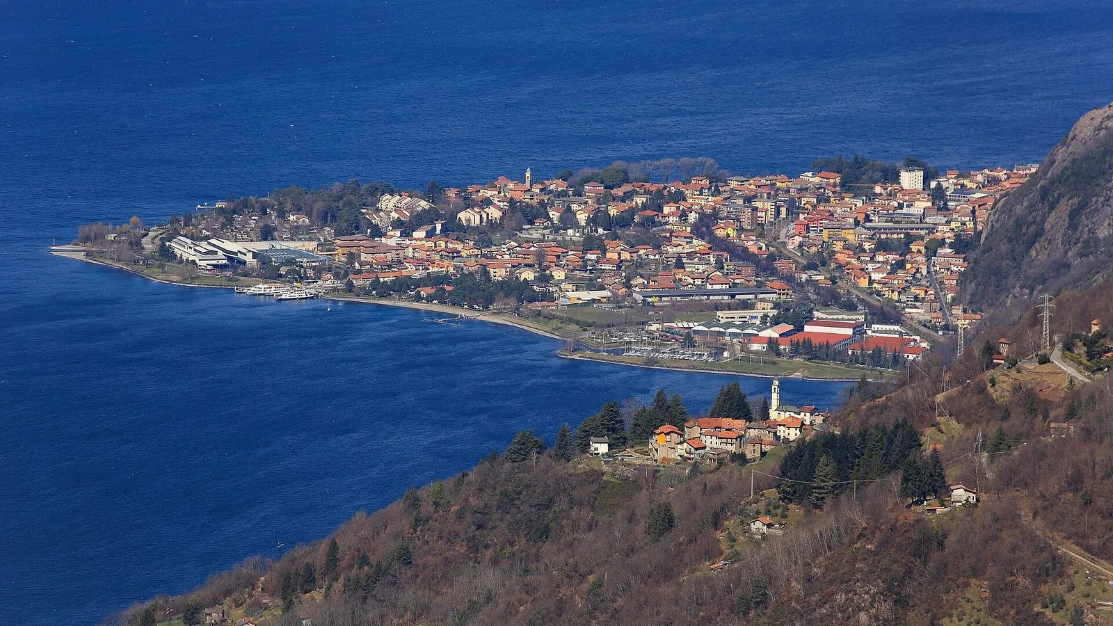 Veduta dall'alto di Dervio sul suo delta che si allunga nel lago blu: le case dai tetti rossi, il porticciolo e la riva, in primo piano un piccolo nucleo con campanile sul pendio boscoso